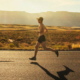Shirtless male runner wearing athletic shorts, running shoes, a hat, and sunglasses while jogging along a road in Arizona
