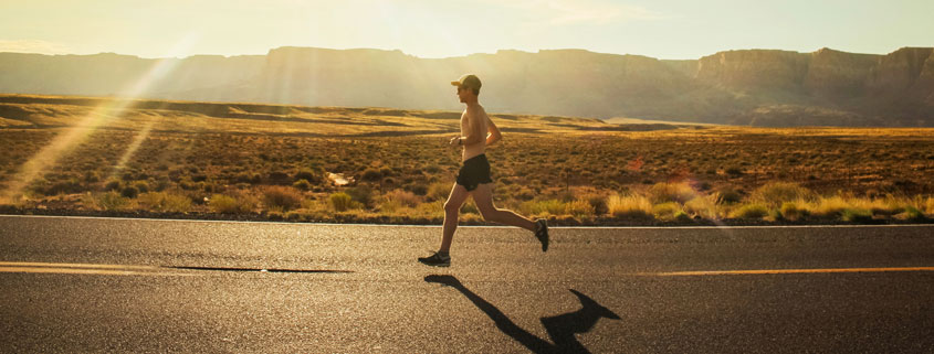 Shirtless male runner wearing athletic shorts, running shoes, a hat, and sunglasses while jogging along a road in Arizona