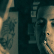 Teenager holding a cigarette to his mouth with two fingers while looking at his reflection in a mirror, with his back visible in the foreground