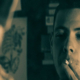 Teenager holding a cigarette to his mouth with two fingers while looking at his reflection in a mirror, with his back visible in the foreground