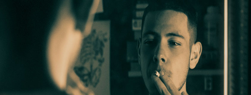 Teenager-Smoking-Cigarette-While-Looking-at-Mirror-Reflection Teenager holding a cigarette to his mouth with two fingers while looking at his reflection in a mirror, with his back visible in the foreground