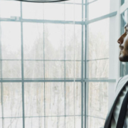 Young businessman wearing a suit standing near an elevator in a modern office building, looking toward a window with a thoughtful expression