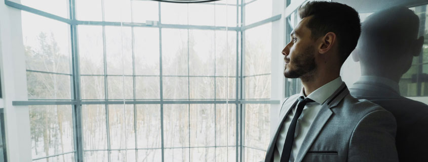 Thoughtful-Businessman-Standing-by-Elevator-in-Modern-Office-Building Young businessman wearing a suit standing near an elevator in a modern office building, looking toward a window with a thoughtful expression