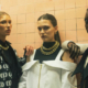 Three women wearing different fashionable outfits standing closely together, with the women on either side resting their arms on the shoulders of the woman in the center