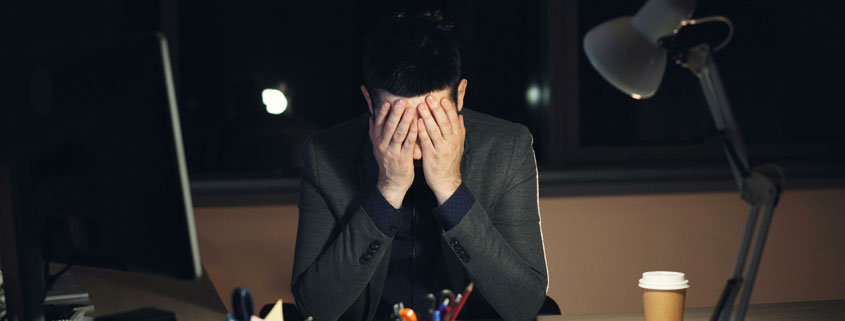 Tired-Office-Worker-Using-Computer-Late-at-Night Tired young businessman wearing a suit and tie working late at night, typing on a computer in a dark office