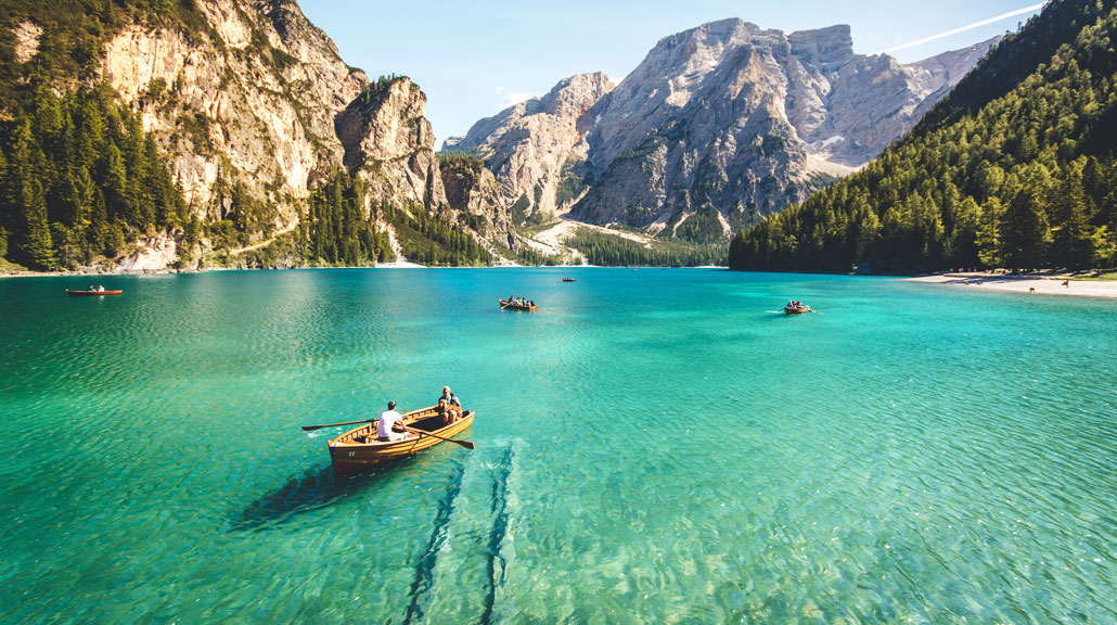 Travelers rowing traditional wooden boats across calm greenish waters surrounded by towering mountains and scenic natural landscapes
