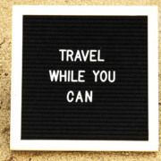 Sign displaying the words Travel While You Can positioned on the sand of a beach