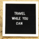 Sign displaying the words Travel While You Can positioned on the sand of a beach