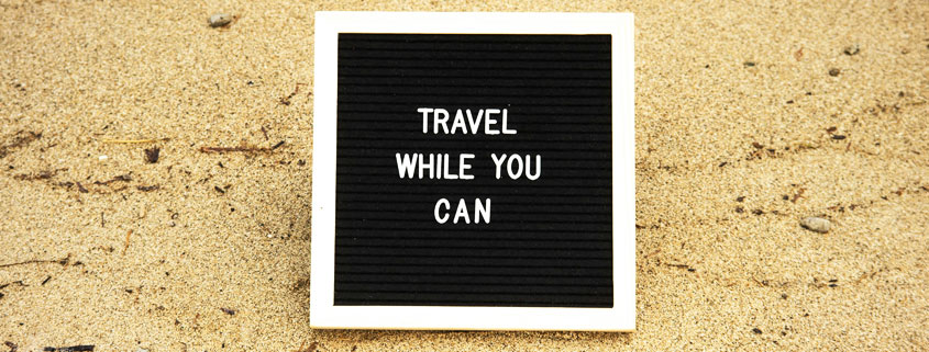 Travel-While-You-Can-Sign-on-Sandy-Beach Sign displaying the words Travel While You Can positioned on the sand of a beach