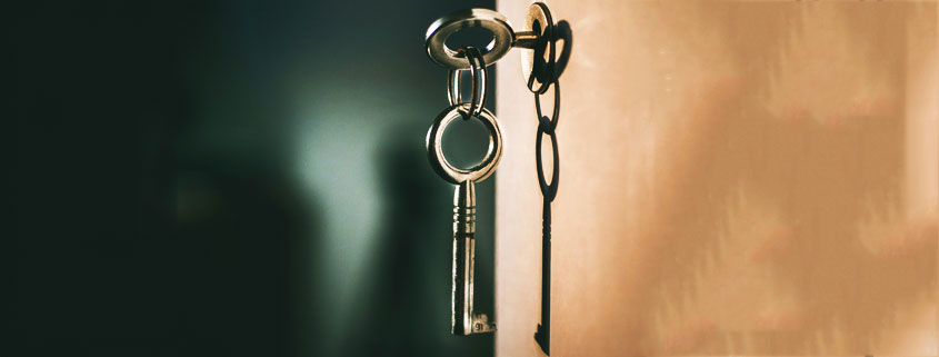Two-Keys-on-Keychain-With-One-Inserted-in-Door-Lock Two metal keys connected by a keychain, with one key inserted into a door lock and the other hanging below