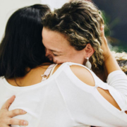 Two women sharing a warm, affectionate hug while a man playing guitar appears softly blurred in the distant background