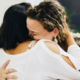 Two women sharing a warm, affectionate hug while a man playing guitar appears softly blurred in the distant background