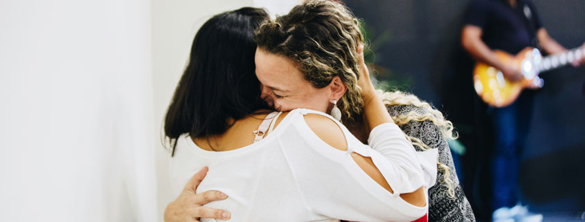 Two-Women-Embracing-in-Warm-Hug-With-Guitarist-in-Background Two women sharing a warm, affectionate hug while a man playing guitar appears softly blurred in the distant background