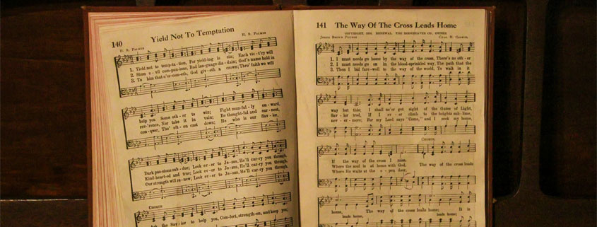 Vintage-Piano-With-Open-Hymn-Book-and-Sheet-Music Open hymn book resting on an old piano displaying sheet music for Yield Not to Temptation and The Way of the Cross Leads Home