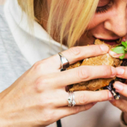 Woman eating a burger or sandwich with a smoothie-style drink beside her, illustrating everyday eating habits and food consumption