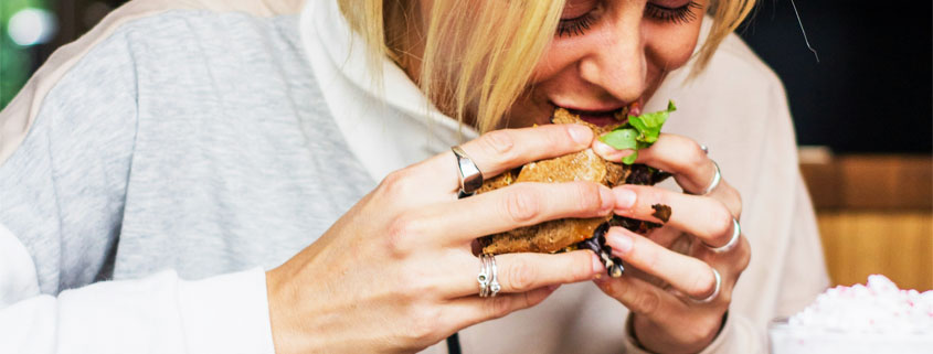 Woman eating a burger or sandwich with a smoothie-style drink beside her, illustrating everyday eating habits and food consumption