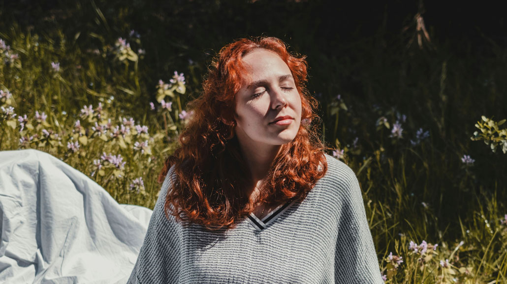 Woman sitting peacefully on a blanket in a grassy field with wildflowers, eyes closed and face lifted toward the warm sunlight
