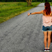 Woman standing on an empty rural road with luggage in one hand and her other arm extended with her thumb raised, appearing to hitchhike