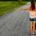 Woman standing on an empty rural road with luggage in one hand and her other arm extended with her thumb raised, appearing to hitchhike