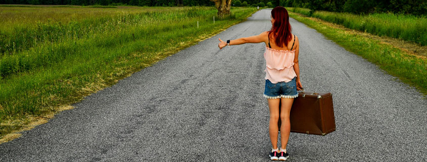 Woman standing on an empty rural road with luggage in one hand and her other arm extended with her thumb raised, appearing to hitchhike