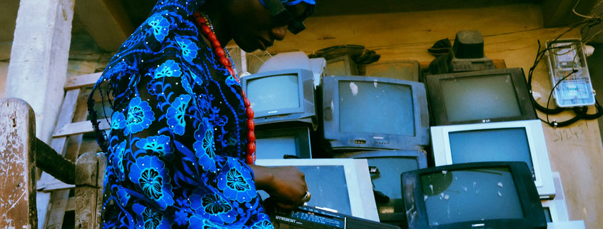 Woman-Holding-Vintage-Cassette-Radio-Surrounded-by-Old-Televisions Black woman holding a vintage cassette radio with a collection of old-fashioned television sets displayed behind her