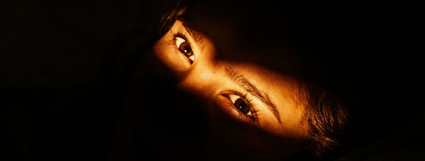 Woman lying in bed in a dark room with a narrow beam of light illuminating her eyes and eyebrows while the rest of her face remains in shadow