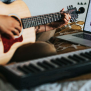 Woman sitting on a bed and playing an acoustic guitar with a piano and laptop positioned in front of her