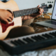 Woman sitting on a bed and playing an acoustic guitar with a piano and laptop positioned in front of her