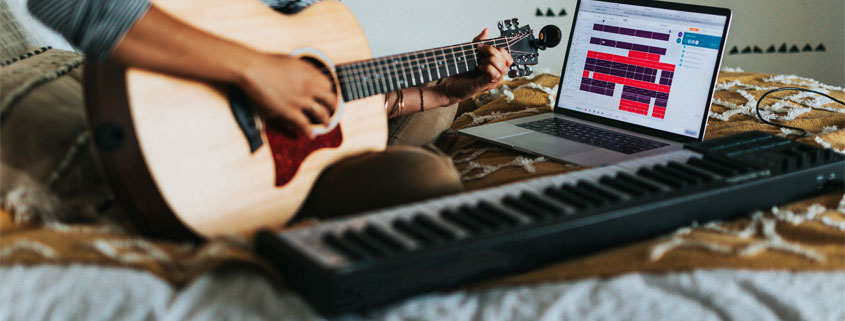 Woman-Playing-Acoustic-Guitar-With-Piano-and-Laptop Woman sitting on a bed and playing an acoustic guitar with a piano and laptop positioned in front of her