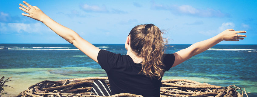 Woman-Relaxing-in-Woven-Basket-Boat-With-Arms-Raised-Near-Shore Woman sitting in a round woven basket boat near the shoreline with both arms raised in the air