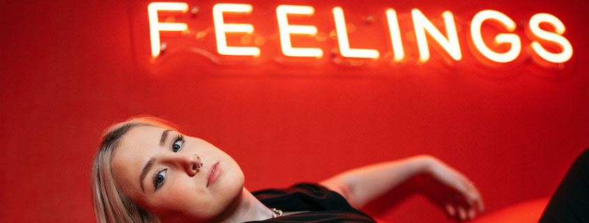 Woman-Relaxing-on-Sofa-With-Feelings-Sign Woman lying comfortably on a sofa with her eyes open and one hand visible beneath a sign displaying the word Feelings