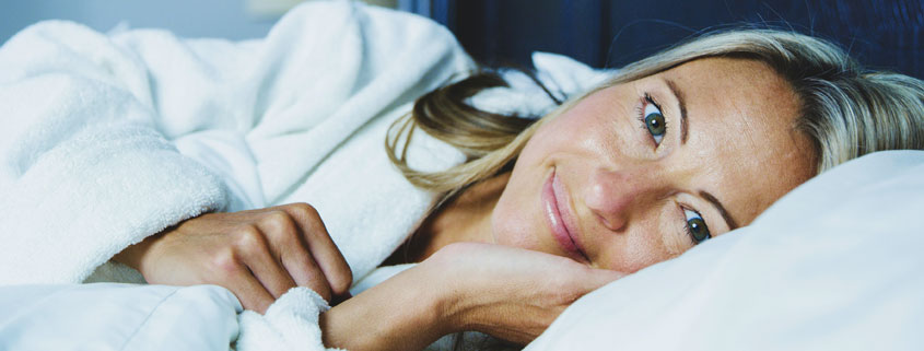 Woman-Resting-on-Bed-Wearing-White-Robe Woman wearing a white robe lying comfortably on a bed while resting