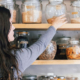 Woman with long hair reaching for a labeled glass jar among shelves filled with neatly organized dry foods and pantry ingredients