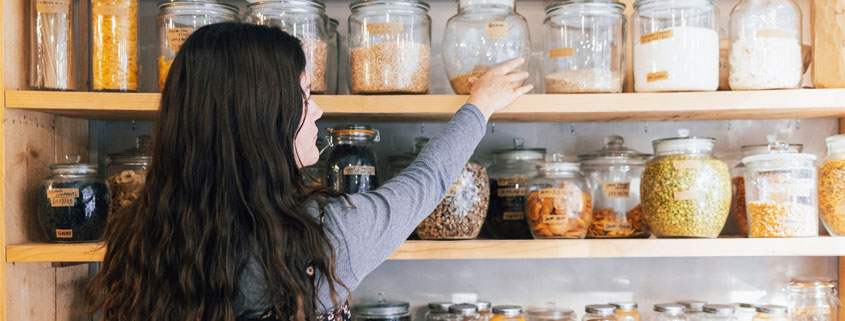 Woman-Selecting-Food-Jar-From-Organized-Pantry Woman with long hair reaching for a labeled glass jar among shelves filled with neatly organized dry foods and pantry ingredients