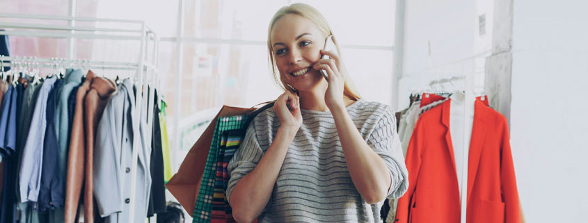 Woman-Shopping-for-Clothes-Talking-on-Phone-in-Fashion-Store Smiling blonde woman carrying shopping bags and talking on her mobile phone while browsing clothing racks in a large fashion store