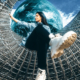 Woman wearing black pants and white boots sitting cross-legged on a curved geometric metal sculpture with a glass-like globe positioned behind her