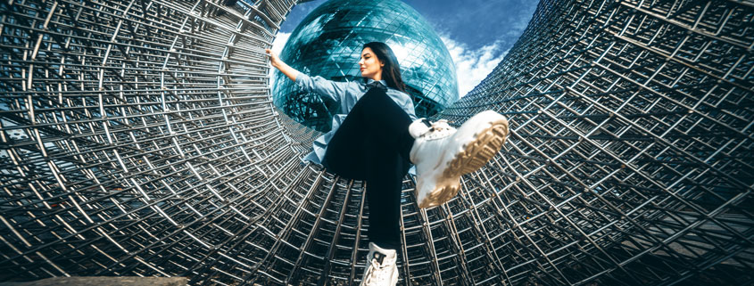 Woman-Sitting-on-Abstract-Metal-Sculpture-With-Glass-Globe-Background Woman wearing black pants and white boots sitting cross-legged on a curved geometric metal sculpture with a glass-like globe positioned behind her