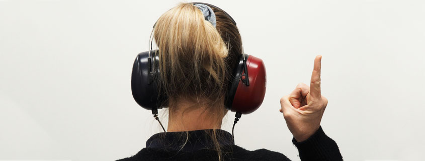 Woman viewed from behind wearing headphones and raising her index finger to indicate hearing a sound during a hearing test