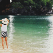 Rear view of a woman standing barefoot at the ocean shoreline, holding a white sun hat with both hands as waves reach her feet