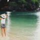 Rear view of a woman standing barefoot at the ocean shoreline, holding a white sun hat with both hands as waves reach her feet