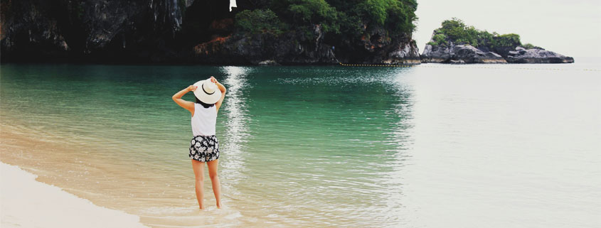 Woman-With-White-Sun-Hat-Standing-at-Ocean-Shore Rear view of a woman standing barefoot at the ocean shoreline, holding a white sun hat with both hands as waves reach her feet