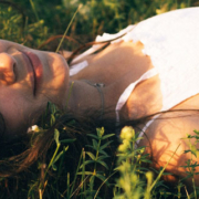 Young girl wearing a white dress resting in a grassy field surrounded by small flowers with a peaceful expression