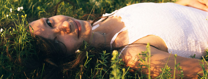 Young-Girl-in-White-Dress-Resting-on-Grass-Among-Flowers Young girl wearing a white dress resting in a grassy field surrounded by small flowers with a peaceful expression