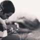 Young boy sitting on grass beside an empty road, concentrating on finger placement while practicing guitar outdoors