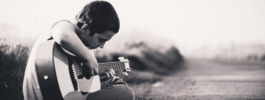 Young boy sitting on grass beside an empty road, concentrating on finger placement while practicing guitar outdoors