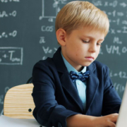 Young student typing on a laptop and writing at a classroom desk with mathematical formulas visible on a blackboard in the background