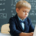 Young student typing on a laptop and writing at a classroom desk with mathematical formulas visible on a blackboard in the background