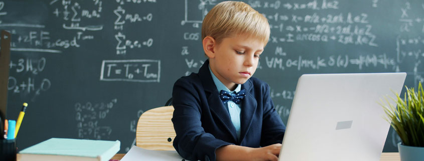 Young-Student-Using-Laptop-and-Writing-in-Classroom Young student typing on a laptop and writing at a classroom desk with mathematical formulas visible on a blackboard in the background