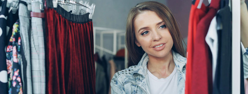Young-Woman-Browsing-Fashionable-Clothes-in-Luxury-Boutique Smiling young woman browsing colorful jumpers, fashionable tops, and skirts while shopping in a luxury clothing boutique