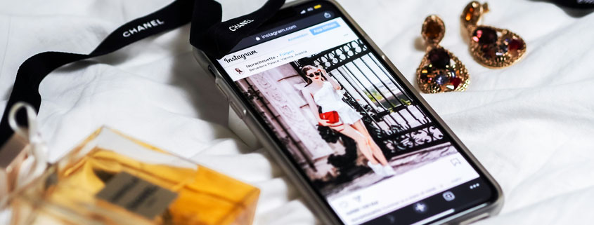 iPhone-With-Chanel-Lanyard-Perfume-and-Elegant-Earrings iPhone resting beside elegant earrings, a perfume bottle, and a black Chanel-branded lanyard arranged on a bed